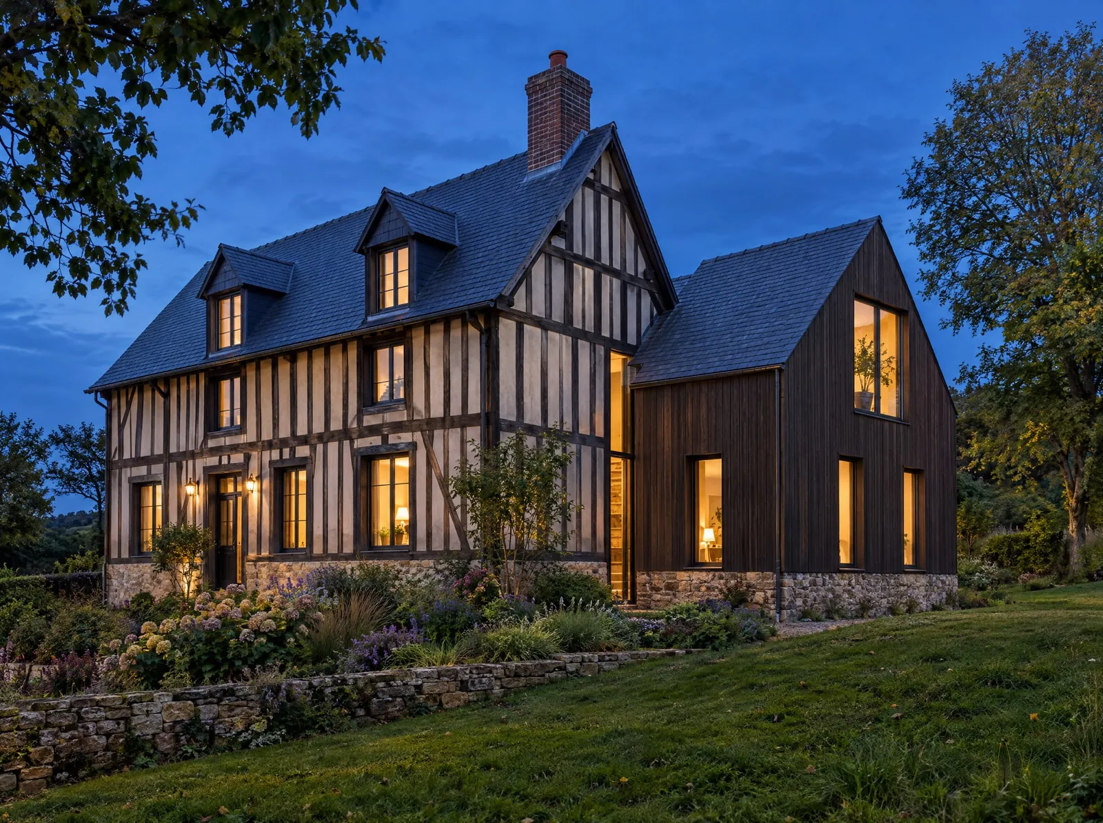 Maison normande à colombages agrandie par un volume à pignon sur deux niveaux, en retrait et de faîtage plus bas, relié par un joint vitré, en bardage bois sombre sur soubassement de pierre.