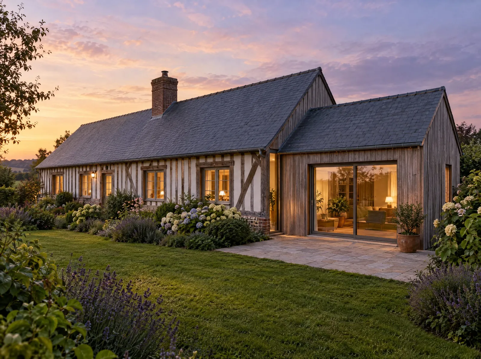 Longère normande à colombages prolongée dans son axe par une extension de plain-pied : même pente de toiture en ardoise, faîtage légèrement abaissé, joint vitré en retrait et bardage de chêne grisé.