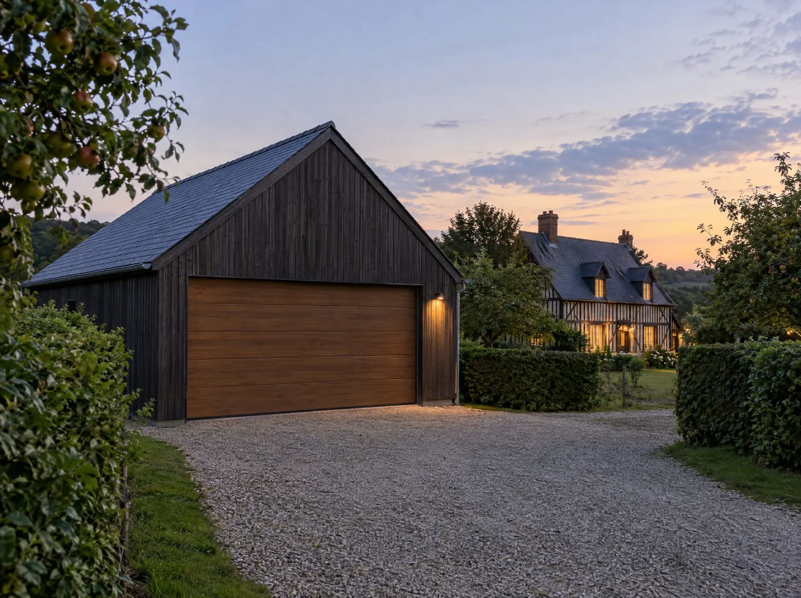 Garage indépendant à bardage bois sombre et toiture en ardoise, devant une maison normande à colombages.